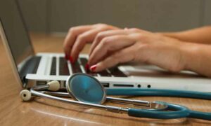 Image of stethoscope next to laptop. Two hands with red nailpolish typing on laptop, Portuguese healthcare for expats | GetNif