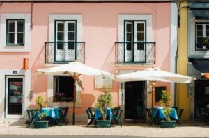 Image of a pink restaurant with green balconies on second floor, on first floor there are three tables and four sets of chairs for El Fresco dining and two umbrellas above the tables for sun protection | Americans eating in Portugal | GetNif