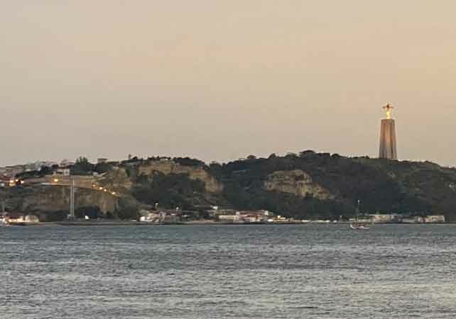 Image of Cristo Rei statute on hill of Lisbon, statue of Christ with arms extended, lit up at dusk, sea underneath him, Portuguese citizenship by investment | GetNif