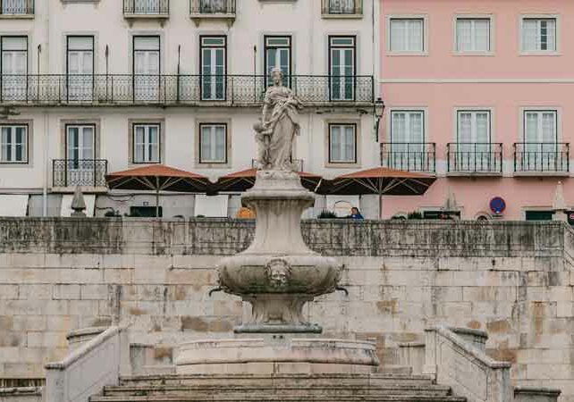 Image of square featuring a marble statue fountain in Lisbon, white building with shading umbrellas out front for diners in background, Golden Visa Requirements | GetNif