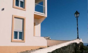 Image of a Portuguese property that is beige and sand in colour featuring large balcony and two windows with shutters, street lamp on corner of house, property owners Portugal | GetNif